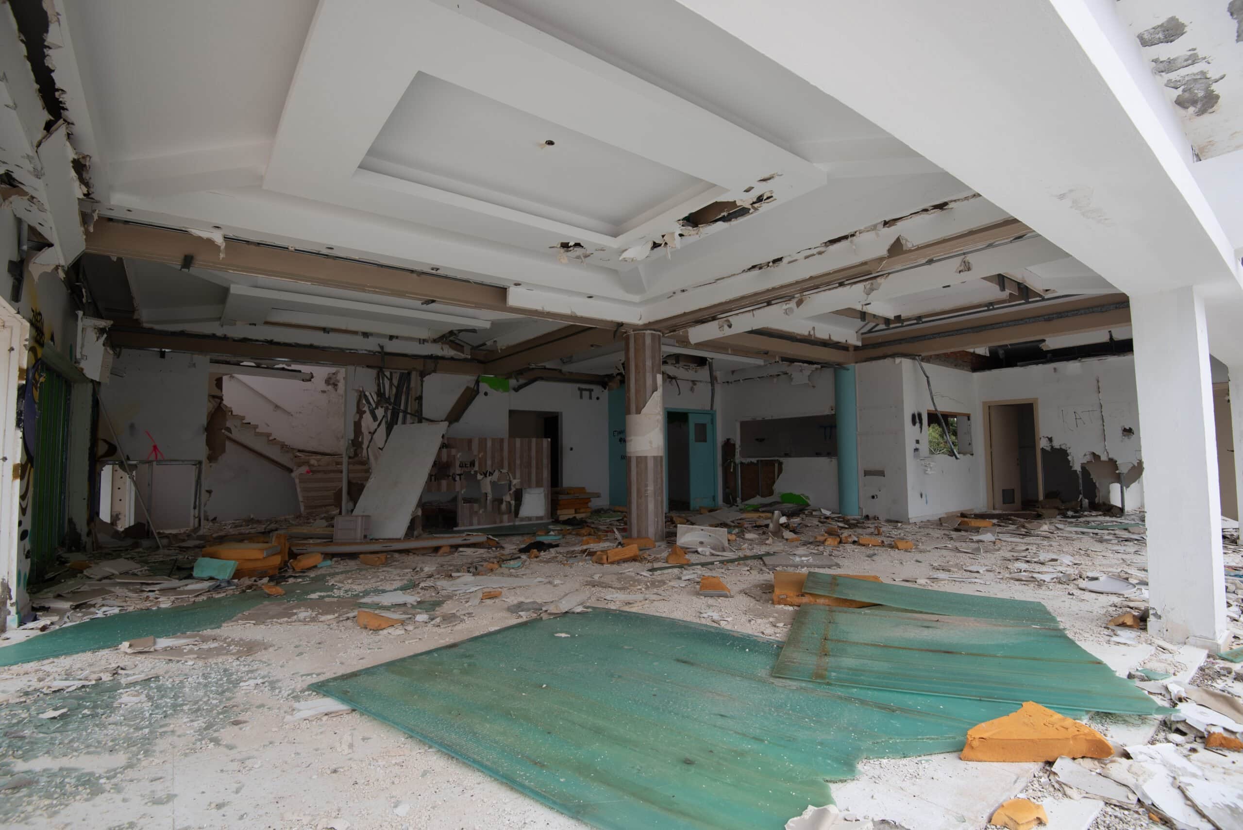 Interior view of a heavily damaged modern building lobby featuring a collapsed tray ceiling, debris-covered floors, and crumbling walls.