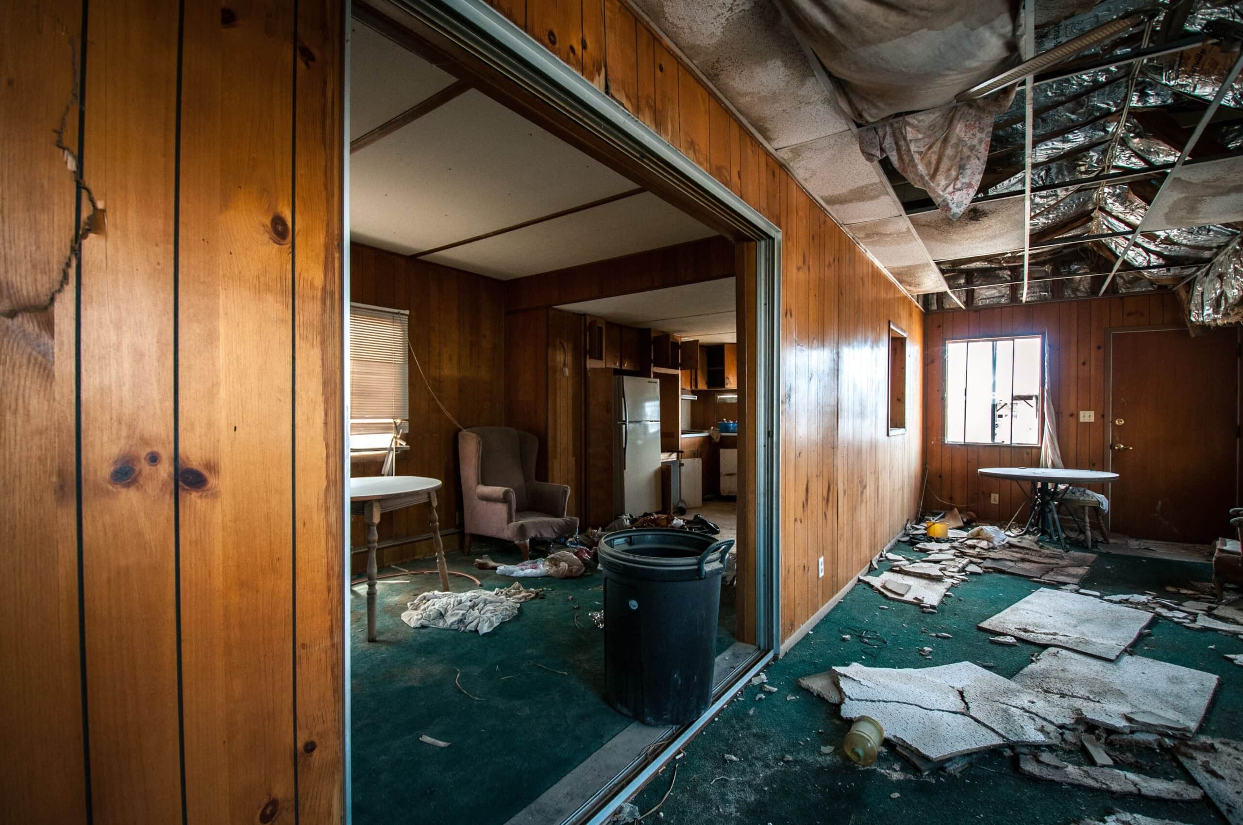 Interior of a neglected home featuring 1970s-style wood-paneled walls, a caved-in ceiling with exposed insulation, and debris scattered across a green carpeted floor.