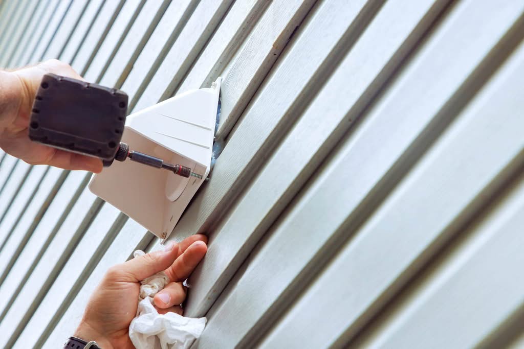 A close-up of a contractor using a power drill to secure a white plastic exterior vent hood onto light gray horizontal vinyl siding.