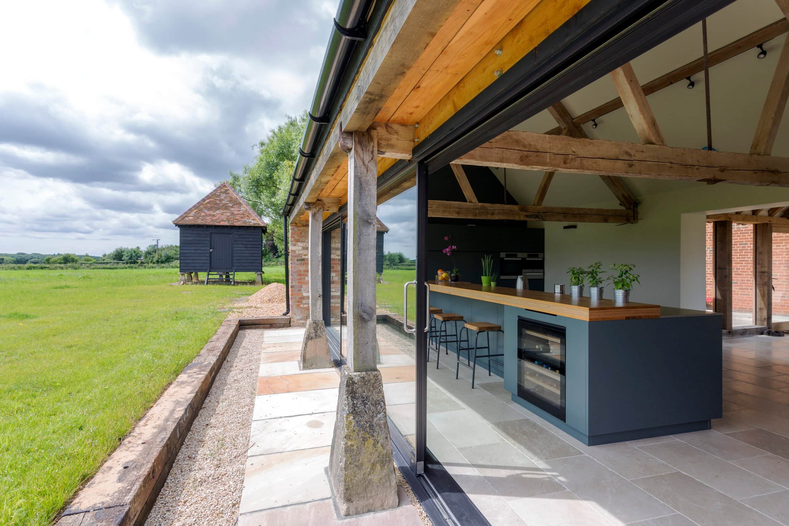 A modern barn conversion featuring an open-plan kitchen with a large teal island, exposed timber beams, and massive sliding glass doors that open to a green countryside view and outdoor living.