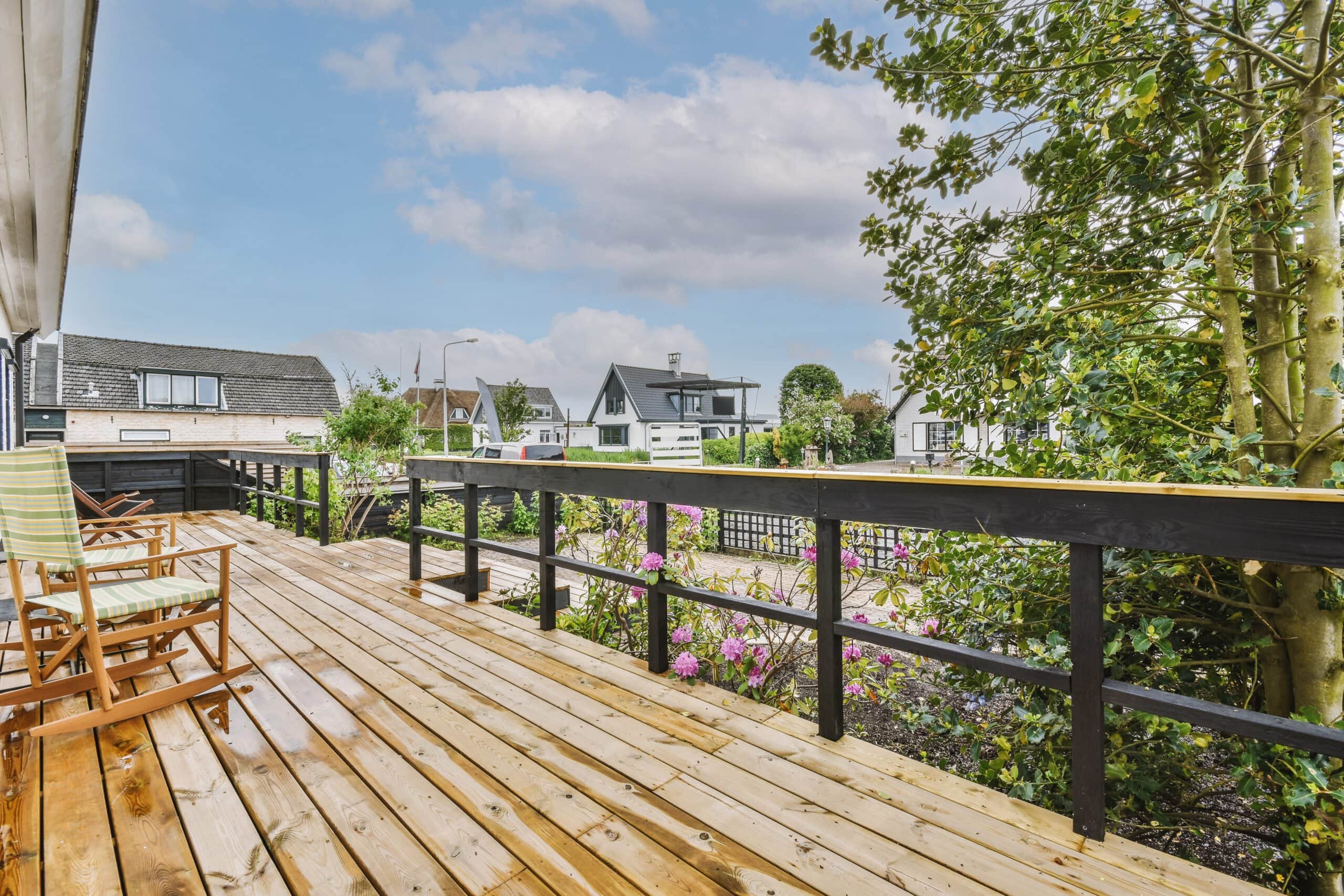 A spacious wooden plank deck with black railings and striped rocking chairs overlooking a coastal residential neighborhood with flowering bushes and trees.