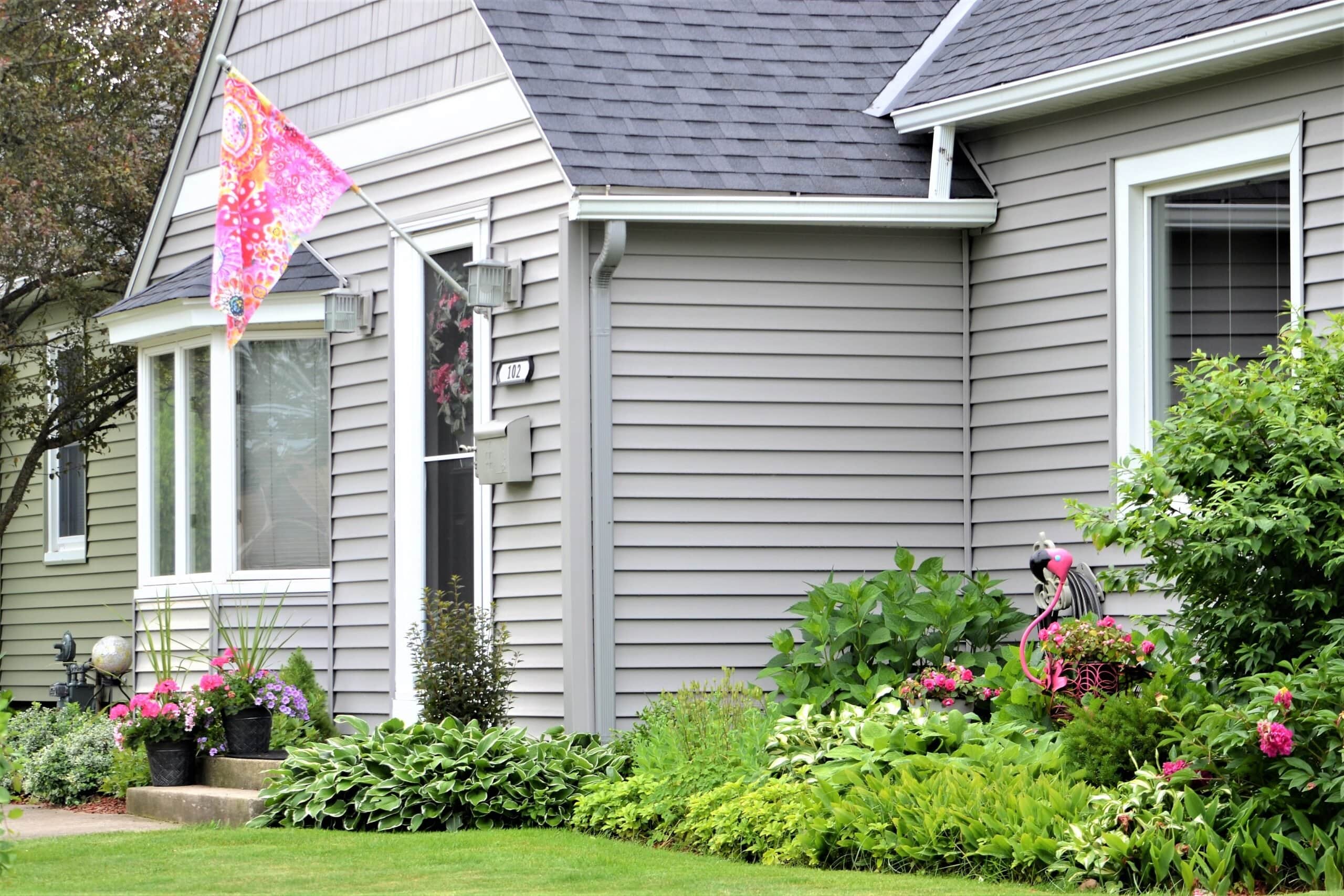 A suburban home featuring light gray horizontal vinyl siding, a dark shingle roof, white window trim, and a vibrant garden with hostas, pink flowers, and a pink flamingo lawn ornament.