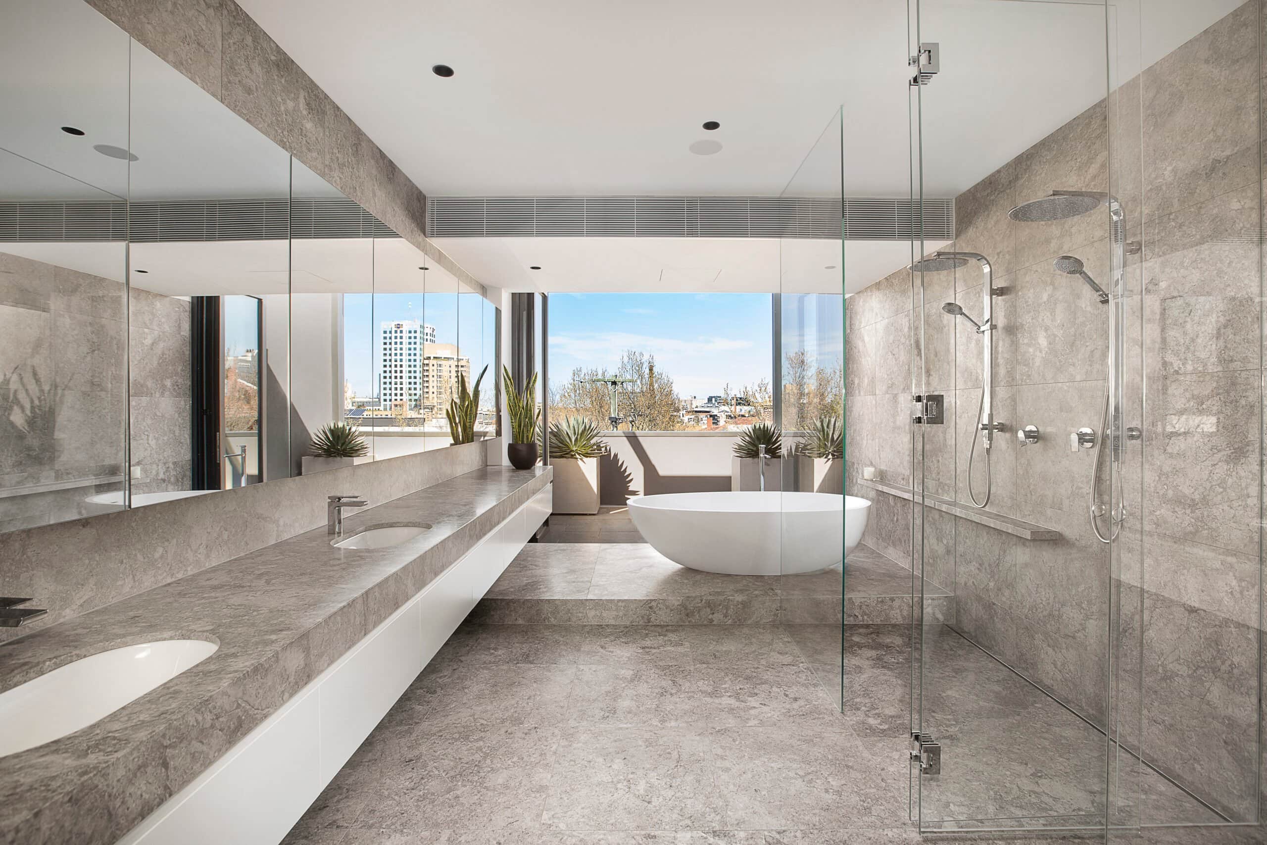 Modern luxury bathroom featuring grey stone tiles, a frameless glass walk-in shower, a freestanding white soaking tub, and a long double vanity with a balcony overlooking a city skyline.