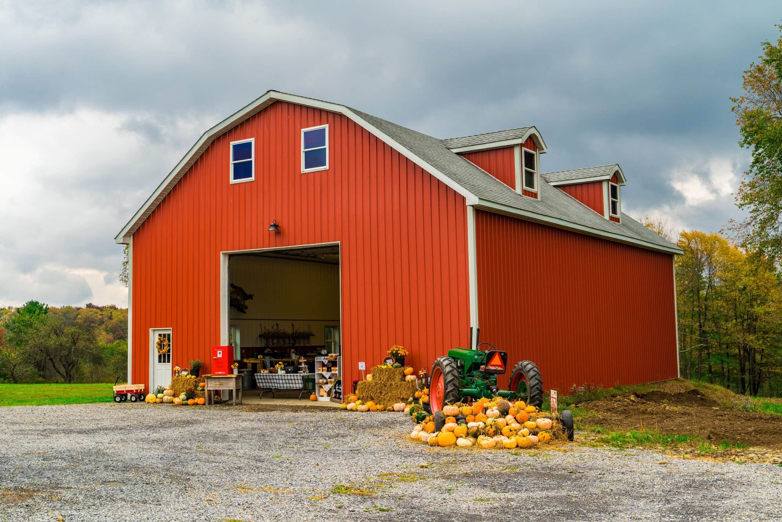 A large, modern red metal barn with a gambrel roof and dormer windows, featuring an open bay door, an autumn display of pumpkins and hay bales, and a green vintage tractor.