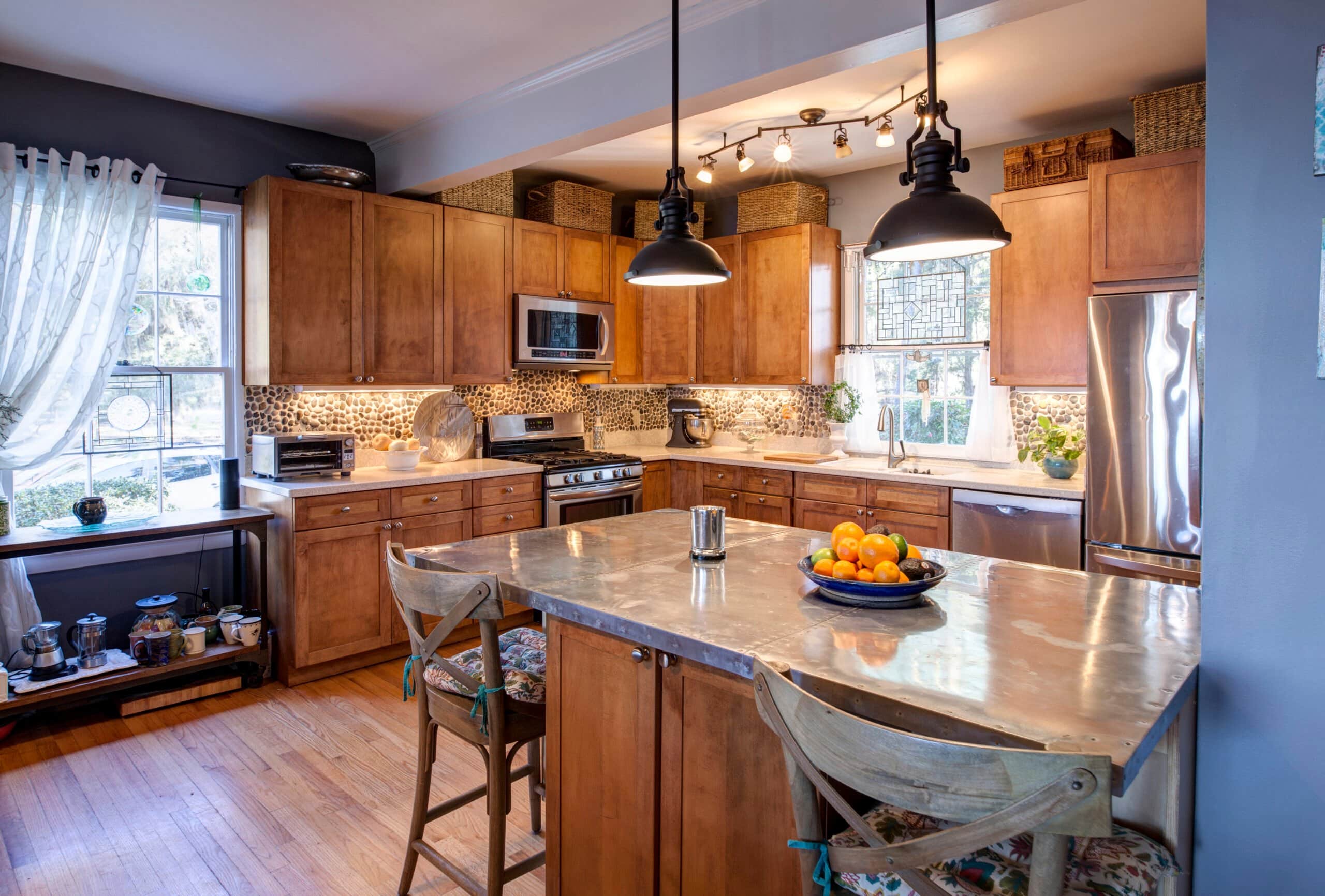 A warm, sunlit kitchen remodeling featuring honey-toned wood cabinets, a large stainless steel-topped island with wooden barstools, and black industrial pendant lights.