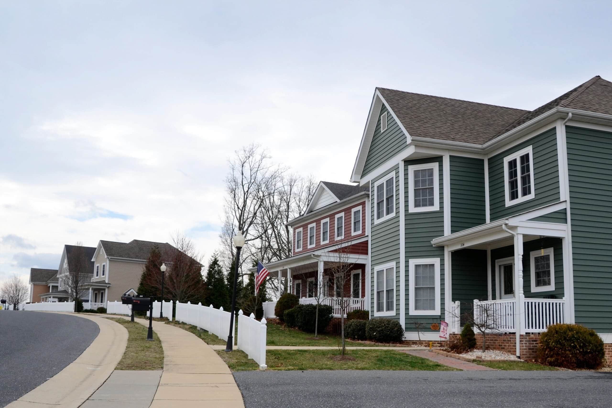 A row of modern two-story suburban homes with various colors of vinyl siding, including forest green, red, and tan, featuring white porches and a white picket fence along a curved sidewalk.