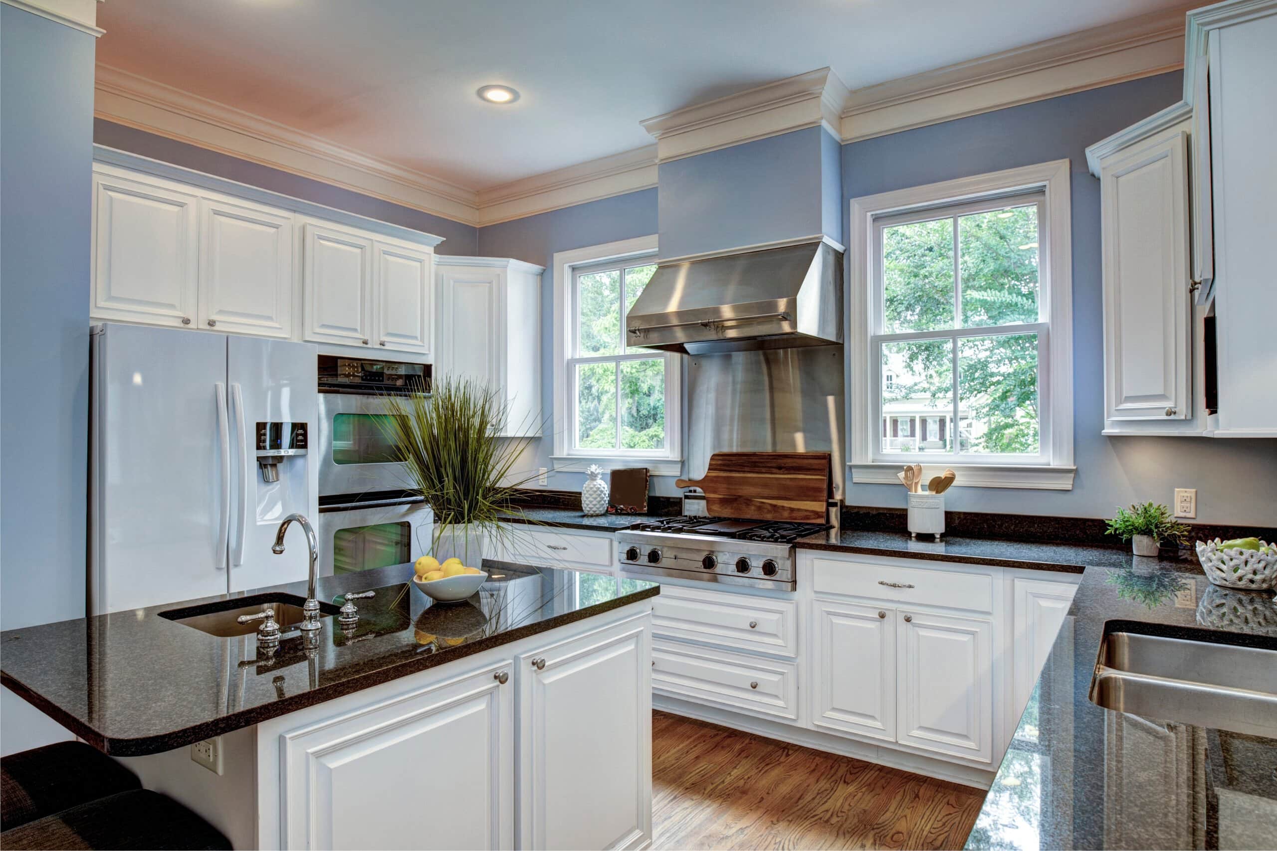 A clean, modern kitchen featuring white shaker cabinets, dark granite countertops, light blue walls, and a professional stainless steel range hood.
