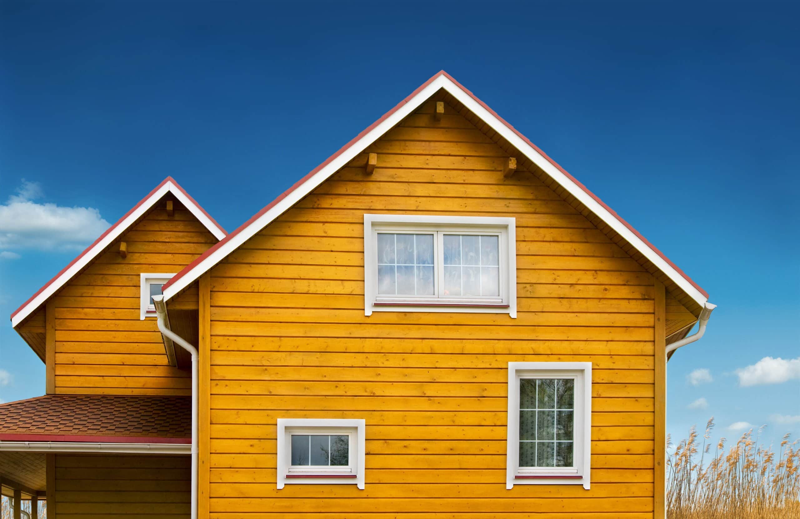 A close-up of a two-story home featuring vibrant yellow horizontal wood-plank siding, white-trimmed windows, and red roof accents under a clear blue sky.