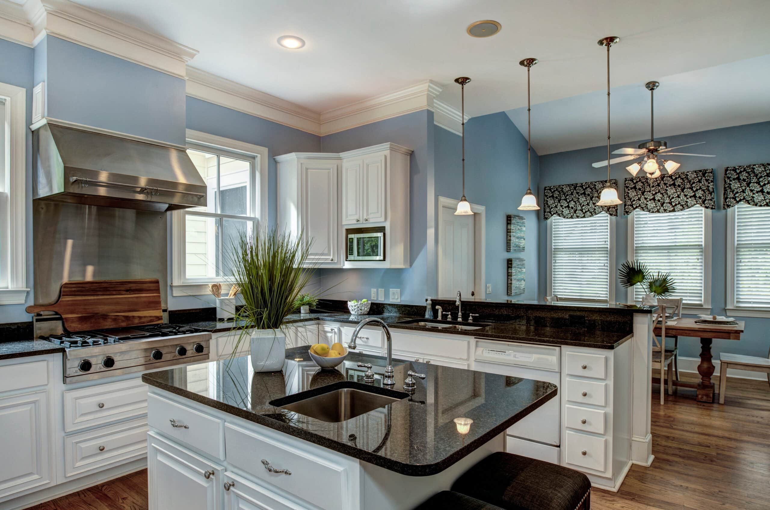 A spacious open-concept kitchen with white cabinetry, dark granite countertops, and an island, leading into a sunlit dining area with blue walls and floral window treatments.