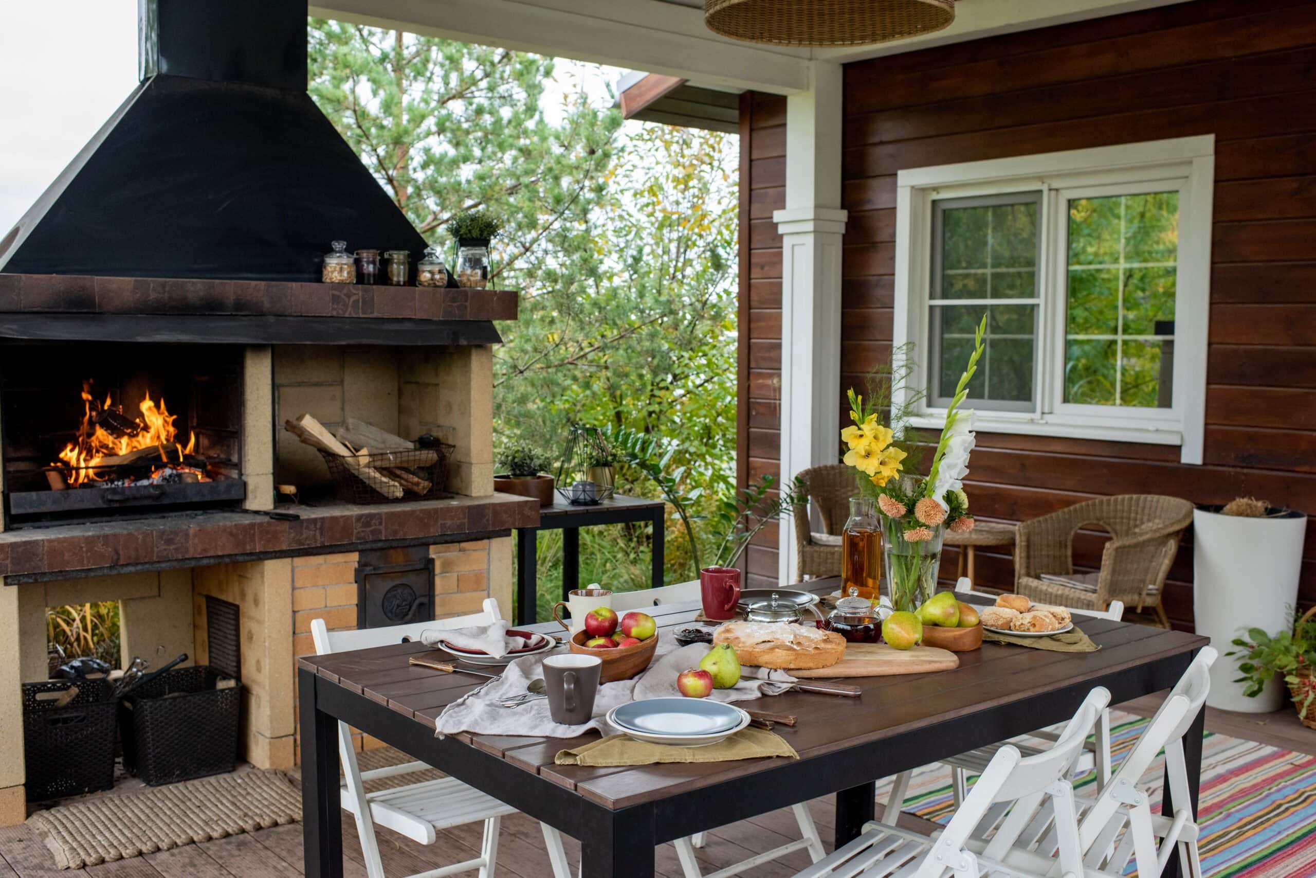 A rustic covered outdoor patio featuring a large brick fireplace with a roaring fire, a wooden dining table set with food and fruit, and white folding chairs against a log cabin wall.