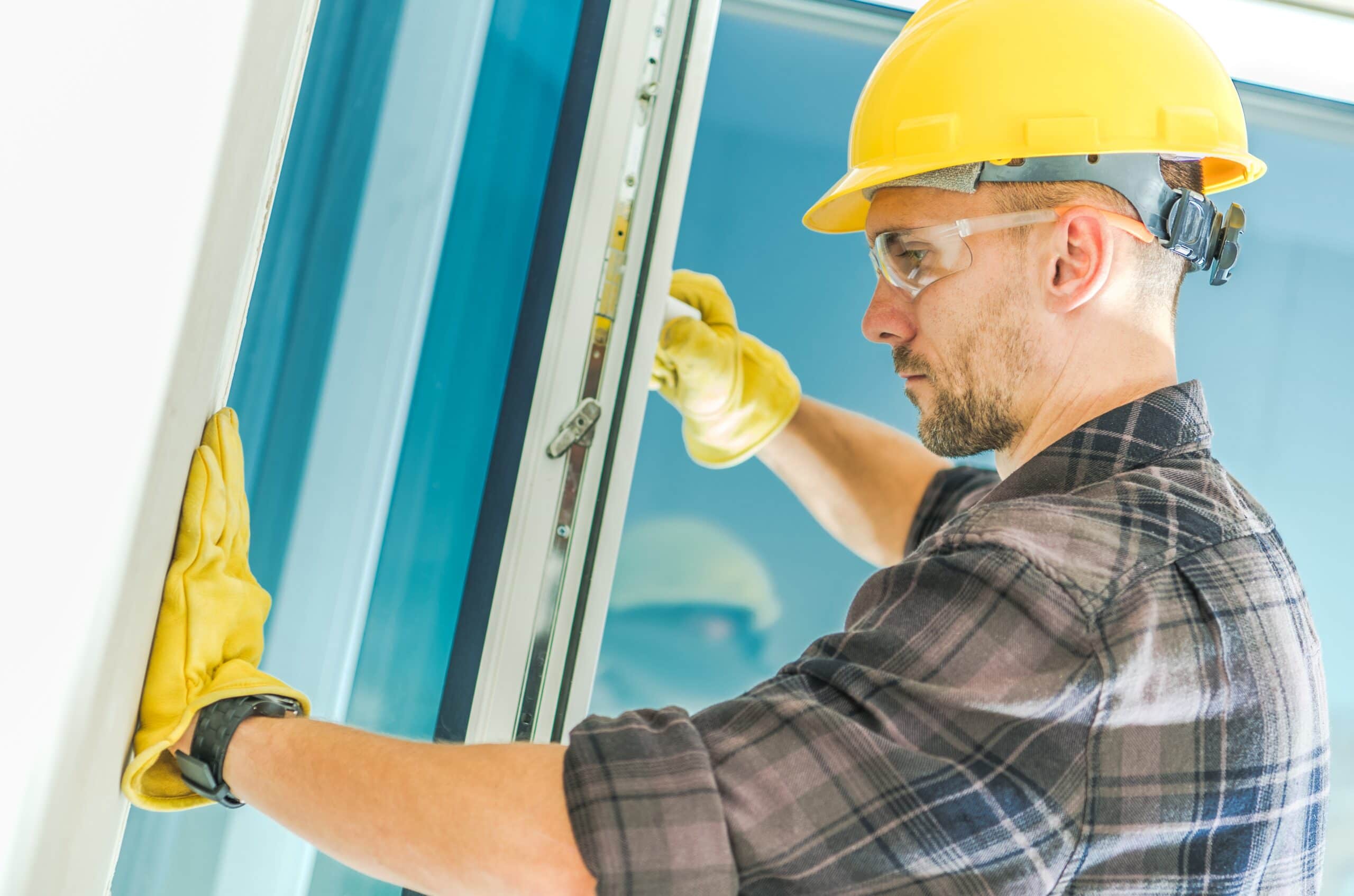 A close-up of a contractor in a yellow hard hat and safety glasses installing a modern white window frame into a residential home.