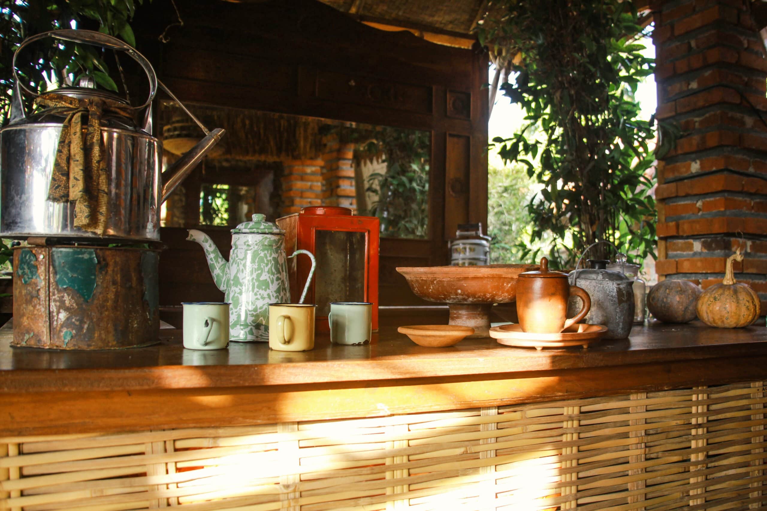 A wooden outdoor counter displaying vintage enamel teapots, colorful mugs, traditional clay pottery, and pumpkins against a bamboo and brick background.