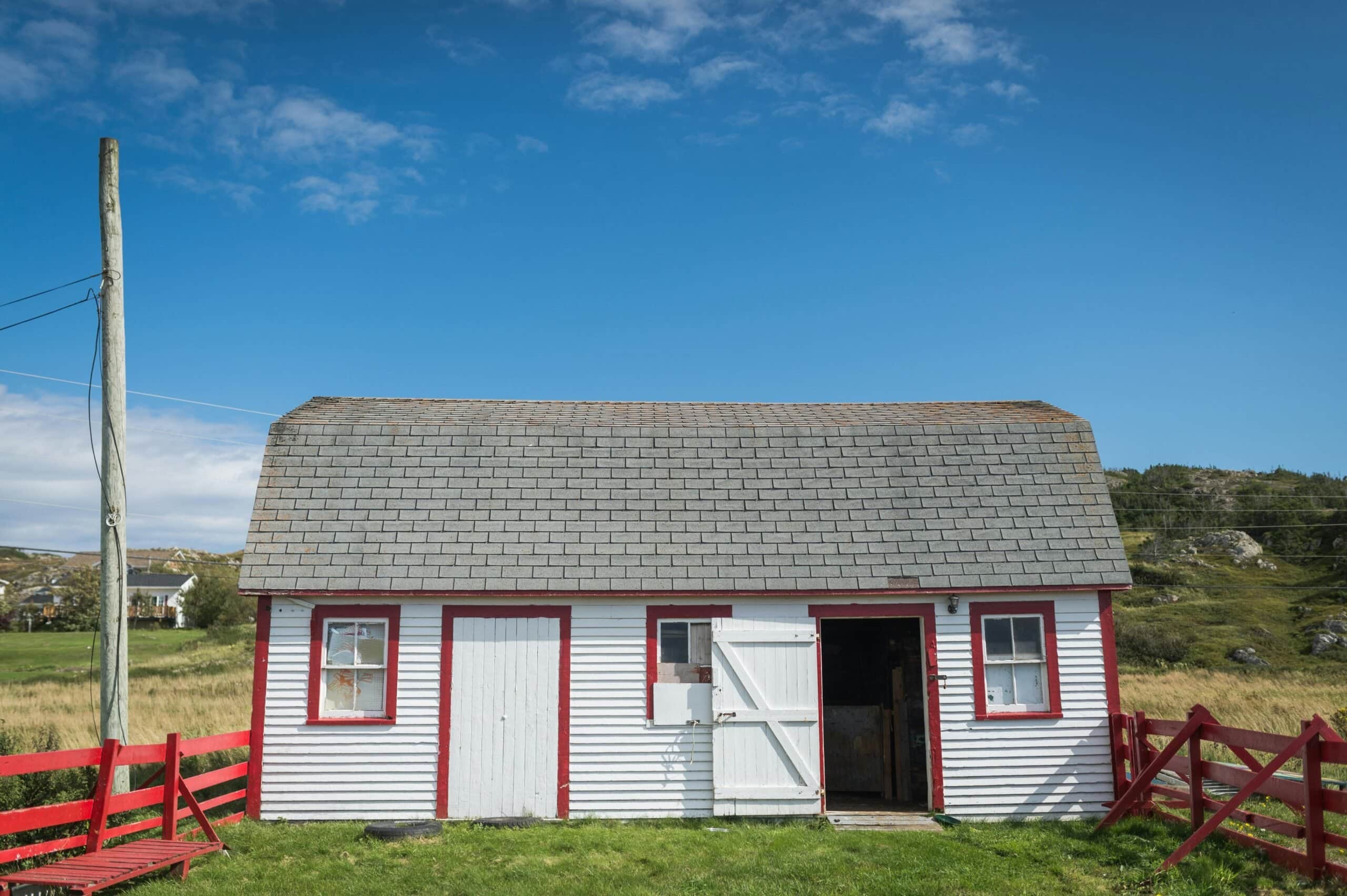 A white horizontal-sided utility shed with red trim around windows and doors, featuring a gray shingle roof and set against a grassy hill under a blue sky.