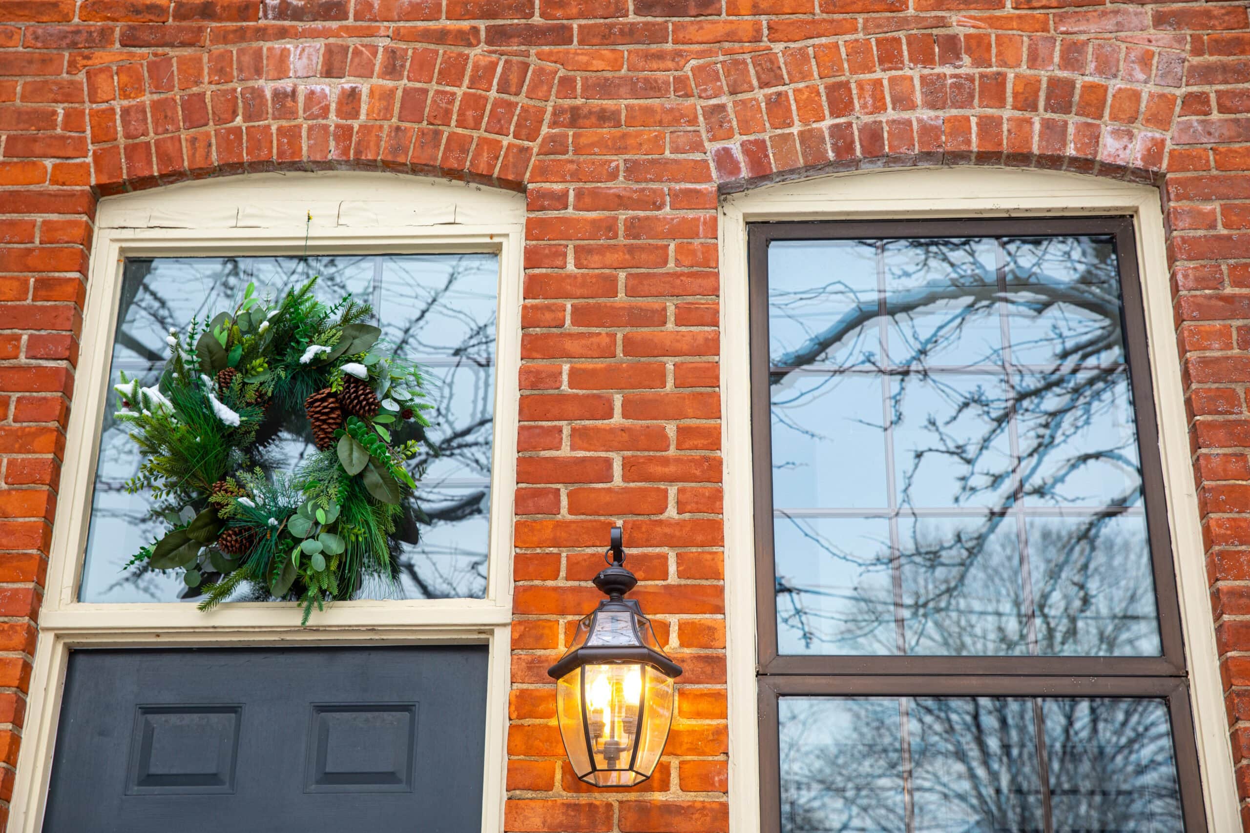 A festive green winter wreath with pinecones hanging in a window above a black door on a red brick building, featuring a glowing outdoor lantern.