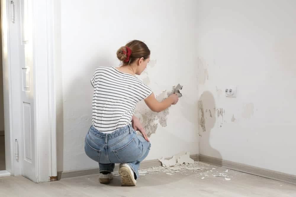 A woman in a striped shirt and jeans kneeling while using a putty knife to scrape peeling paint and plaster from a white interior wall.