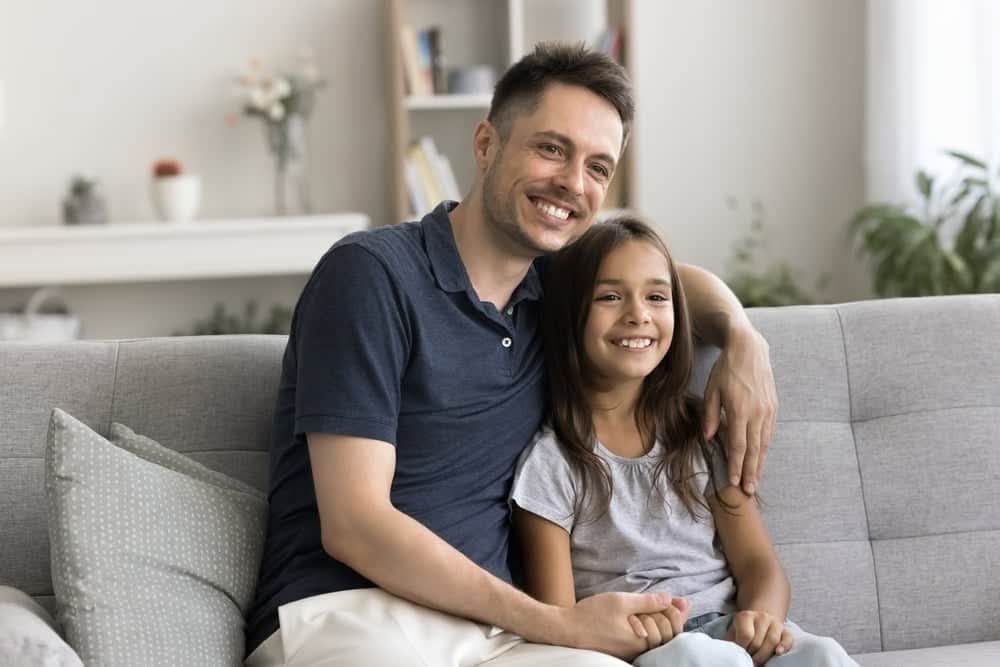 A smiling father and daughter sitting comfortably on a grey sofa in a brightly lit, modern living room after a successful home renovation.