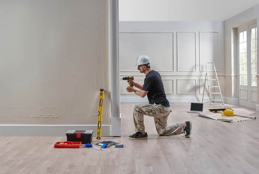 Male contractor in a white hard hat kneeling and using a power drill on a wall during an interior home renovation project.