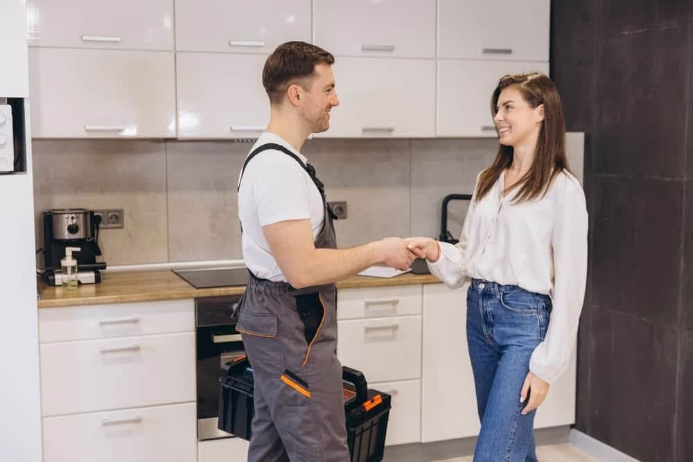 A male handyman in grey work overalls holding a black toolbox shaking hands with a smiling female client in a modern white kitchen.