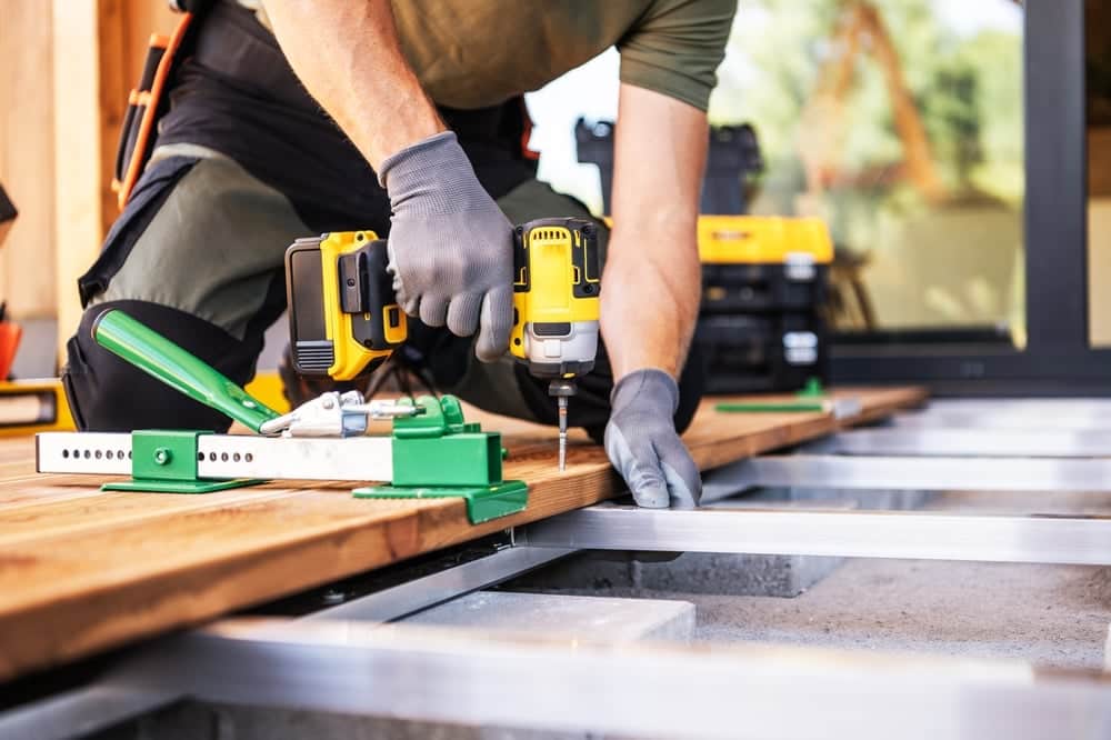 A contractor in work gear kneeling on a wooden deck, using a yellow cordless impact driver and a green board-straightening tool to secure deck planks. Deck Needs Professional Repair.