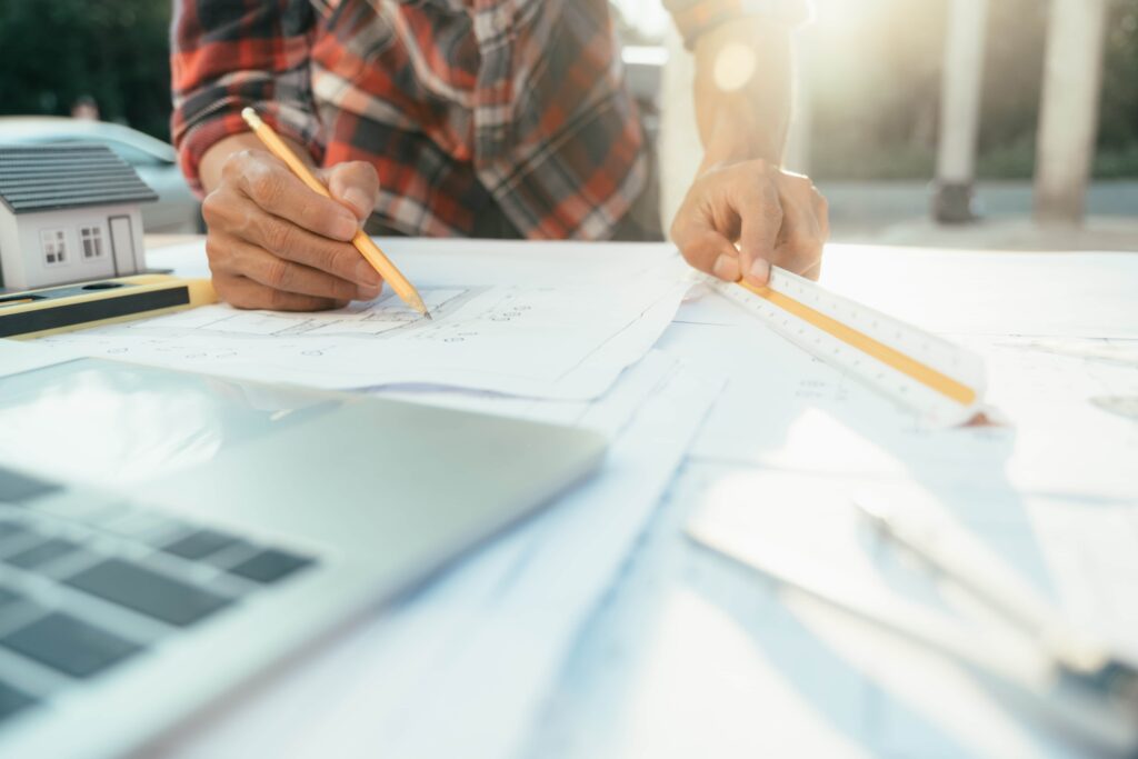 Close-up of an architect and engineer working on residential house blueprints with a pencil, scale ruler, and model home addition on a desk during sunset.