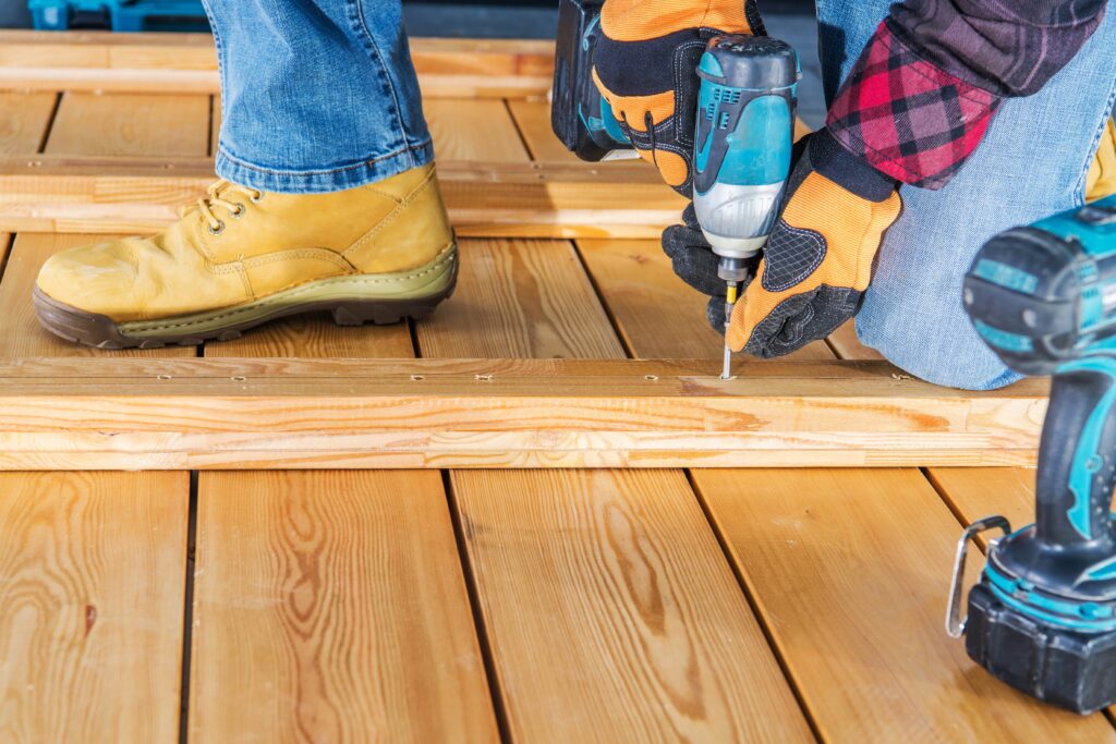 A person wearing work gloves and safety boots using a cordless impact driver to screw a wooden plank onto a deck frame to be the best custom deck builders in the Villages, Florida.