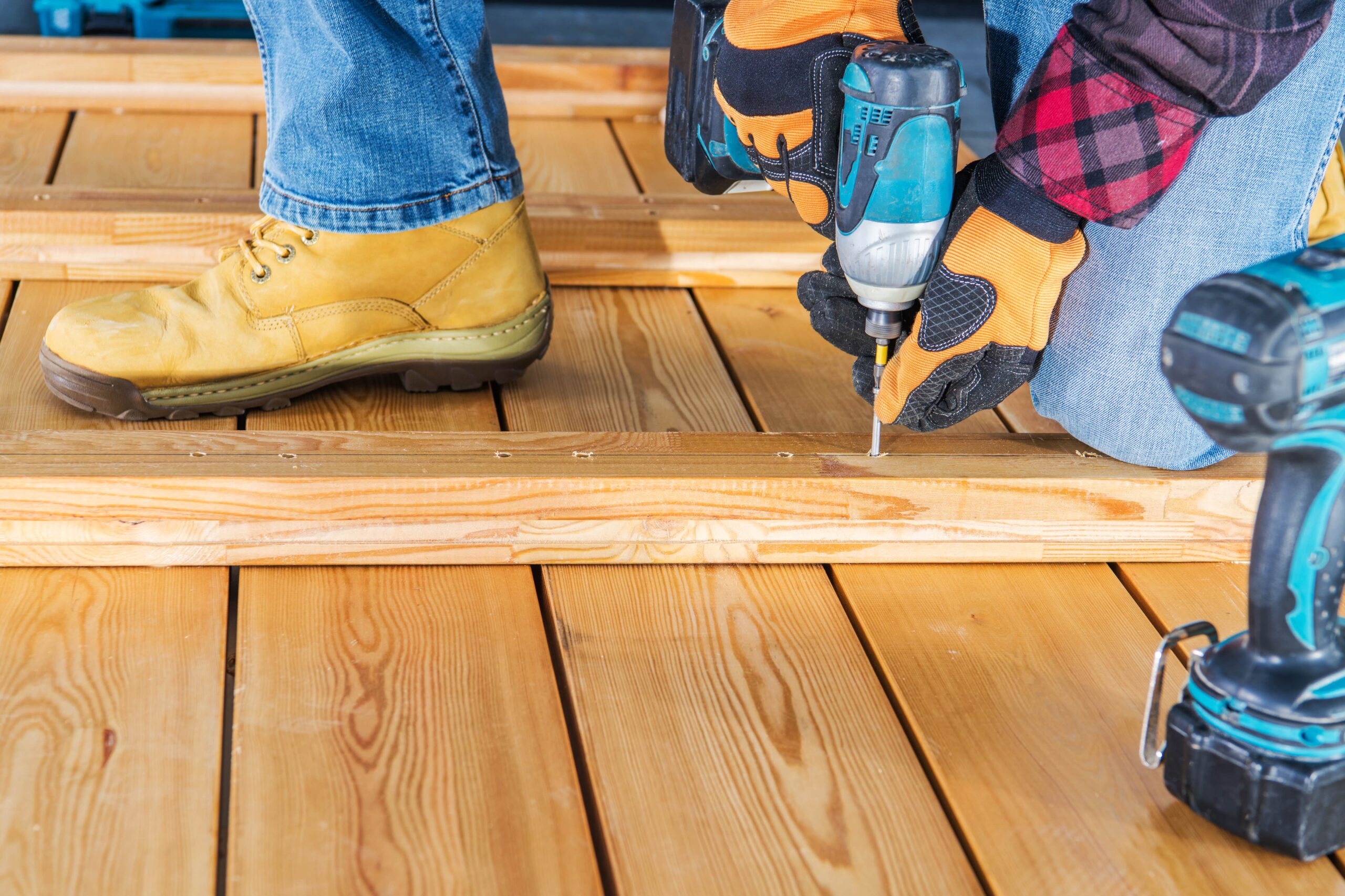 A person wearing work gloves and safety boots using a cordless impact driver to screw a wooden plank onto a deck frame.
