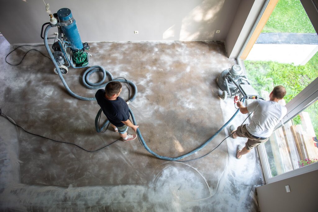 Overhead view of two contractors using a professional floor grinder and a dust extraction vacuum system on a concrete floor in a residential space for water damage restoration in the Villages, Florida.