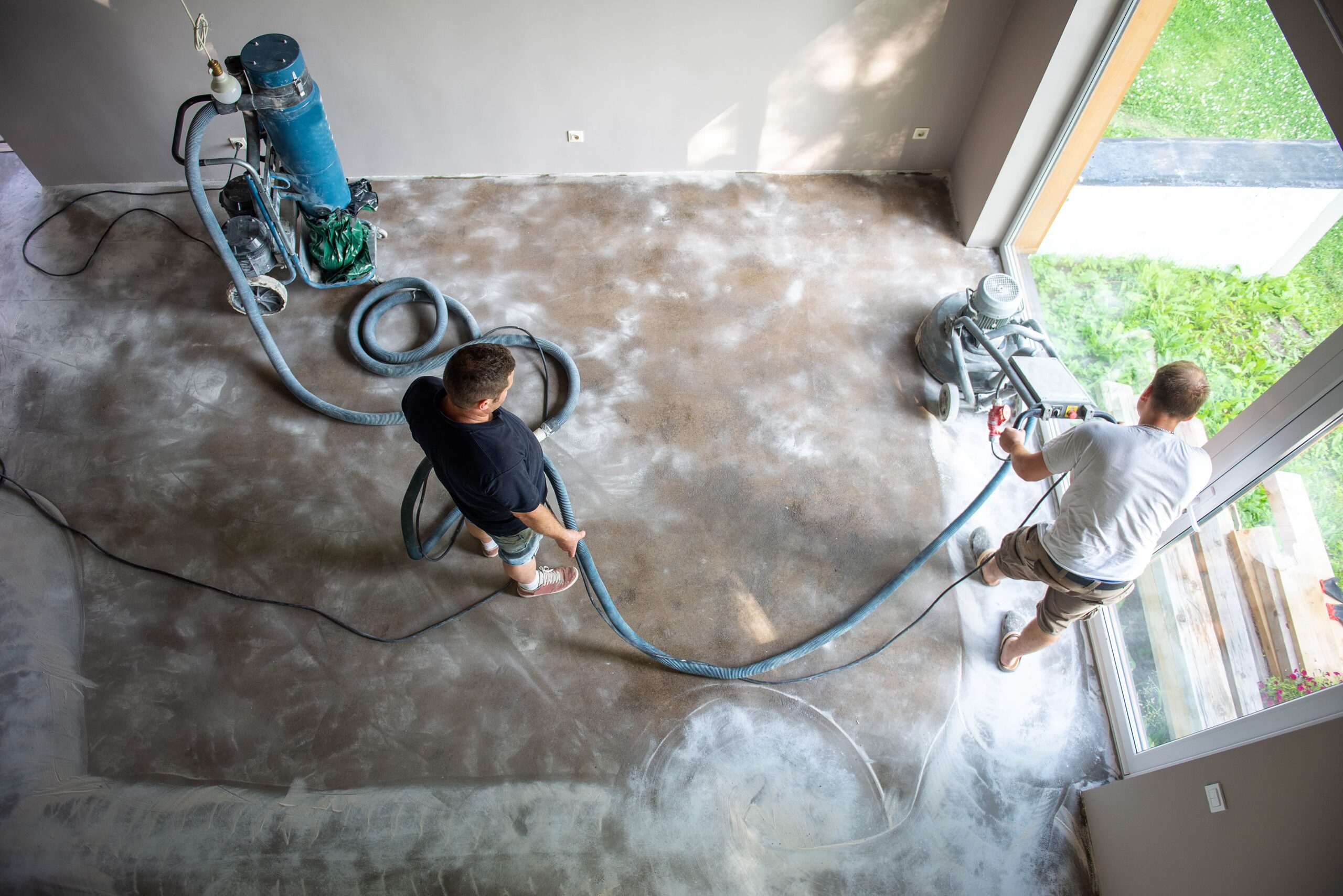 Overhead view of two contractors using a professional floor grinder and a dust extraction vacuum system on a concrete floor in a residential space.