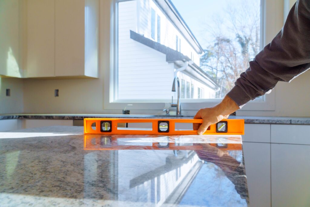 Close-up of a contractor's hand using an orange spirit level on a polished granite flood-proof kitchen island countertop during installation, with a bright window and white kitchen cabinetry in the background.