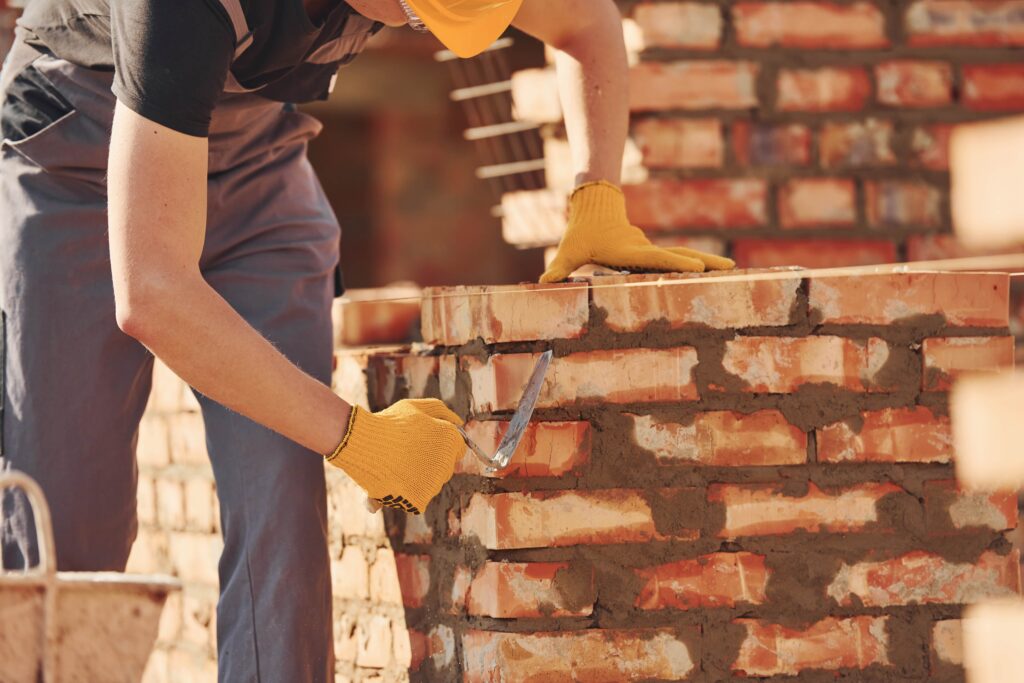 Construction worker in a yellow hard hat and safety gloves using a trowel to apply mortar while laying a red brick wall on a sunny building site. Brick and Stone Veneer Repair Services.