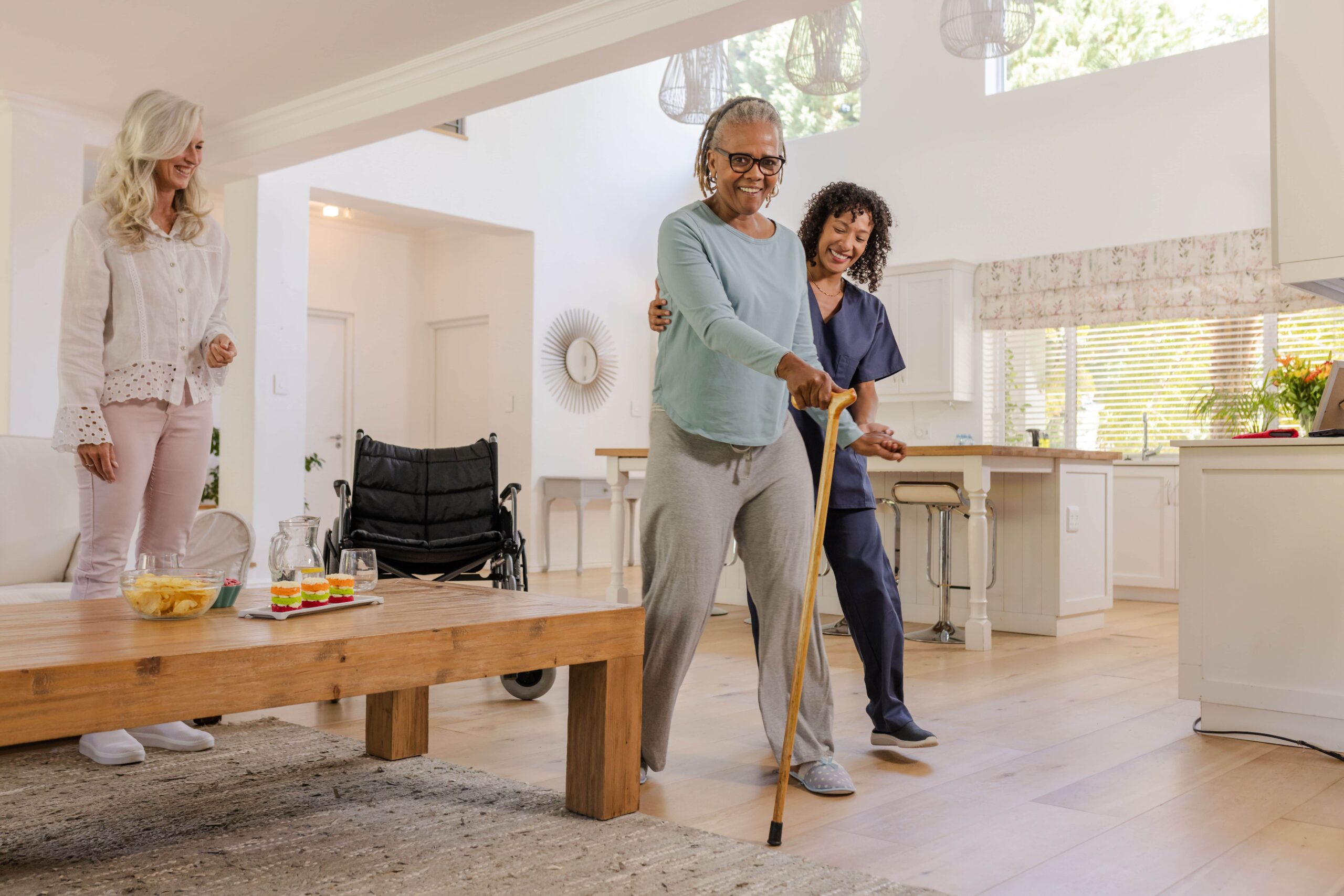 A smiling senior woman uses a wooden walking cane to navigate a bright living room, supported by a professional caregiver in blue scrubs while another woman observes encouragingly; a wheelchair is visible in the background.