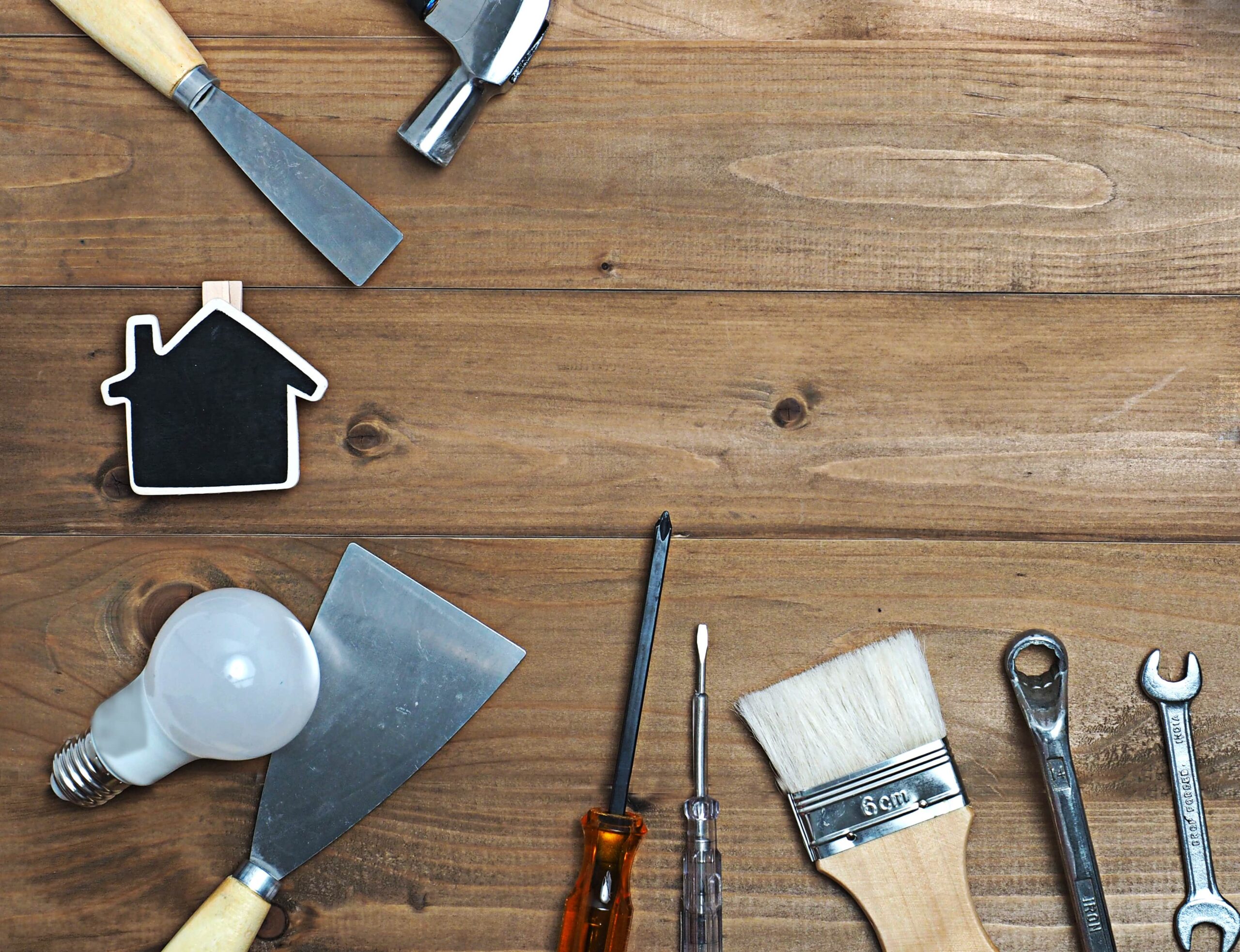 A top-down view of various hand tools including a paintbrush, screwdrivers, wrenches, a putty knife, and a lightbulb arranged on a wooden surface around a small house-shaped chalkboard.