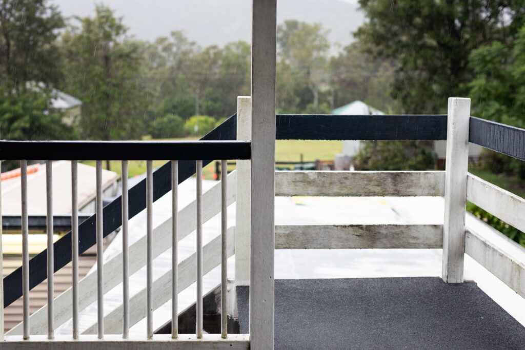 Deck Needs Professional Repair? 5 Signs Before Hurricane Season 3 View from a wooden balcony during rain in Ocala, Florida , showing weathered white and black railings in need of maintenance with a green rural landscape in the background.