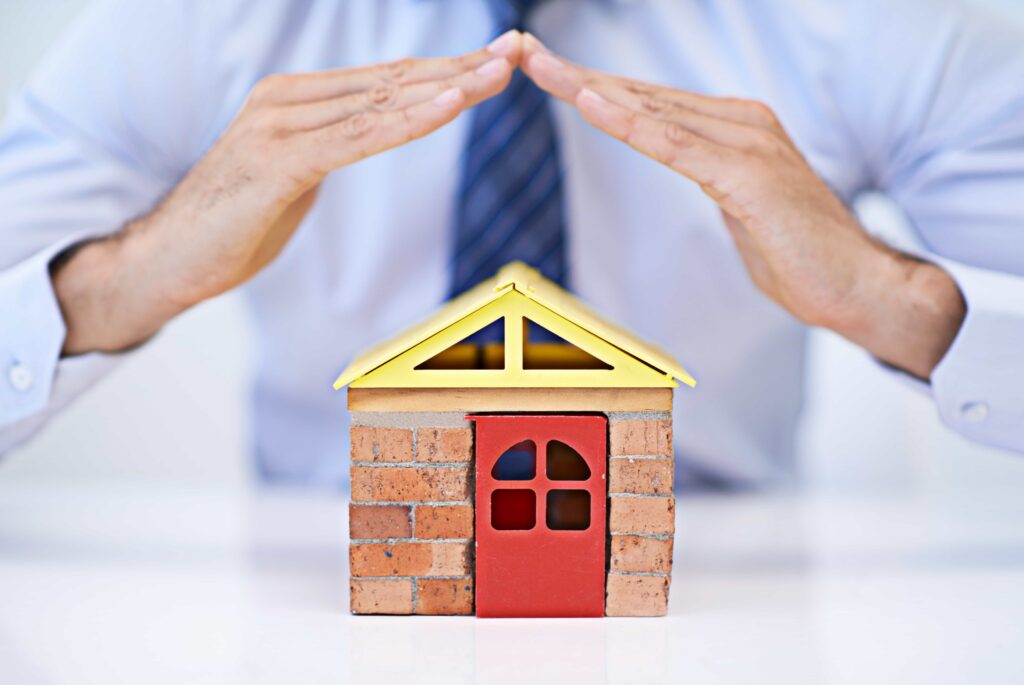 A man in professional attire protecting a small model house with his hands, symbolizing residential asset protection. Expert home safety audit service in Jacksonville, Florida.