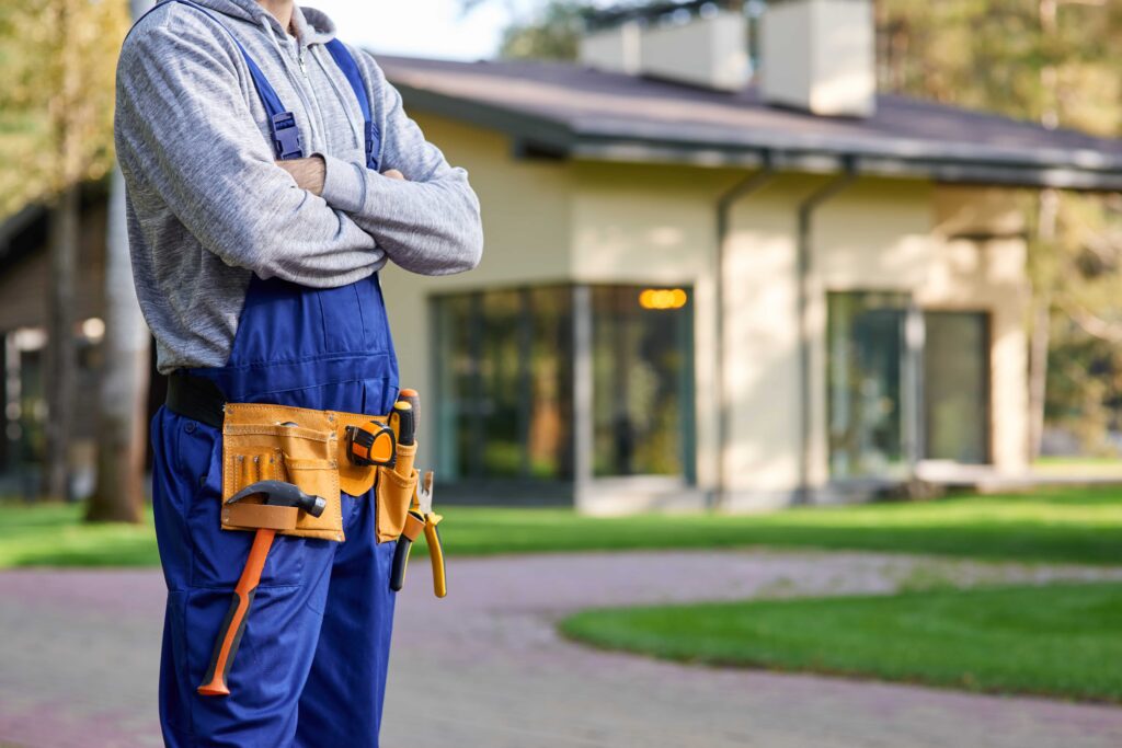 A cropped shot of a professional home renovation contractor with crossed arms wearing blue overalls and a tool belt, standing in front of a preventative home maintenance in Tampa, Florida