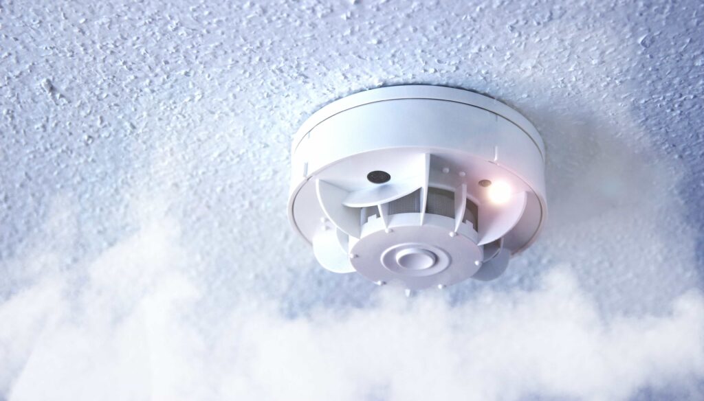 A white smoke detector mounted on a textured ceiling with visible smoke and an active indicator light. Professional residential fire safety service in Tampa, FL.
