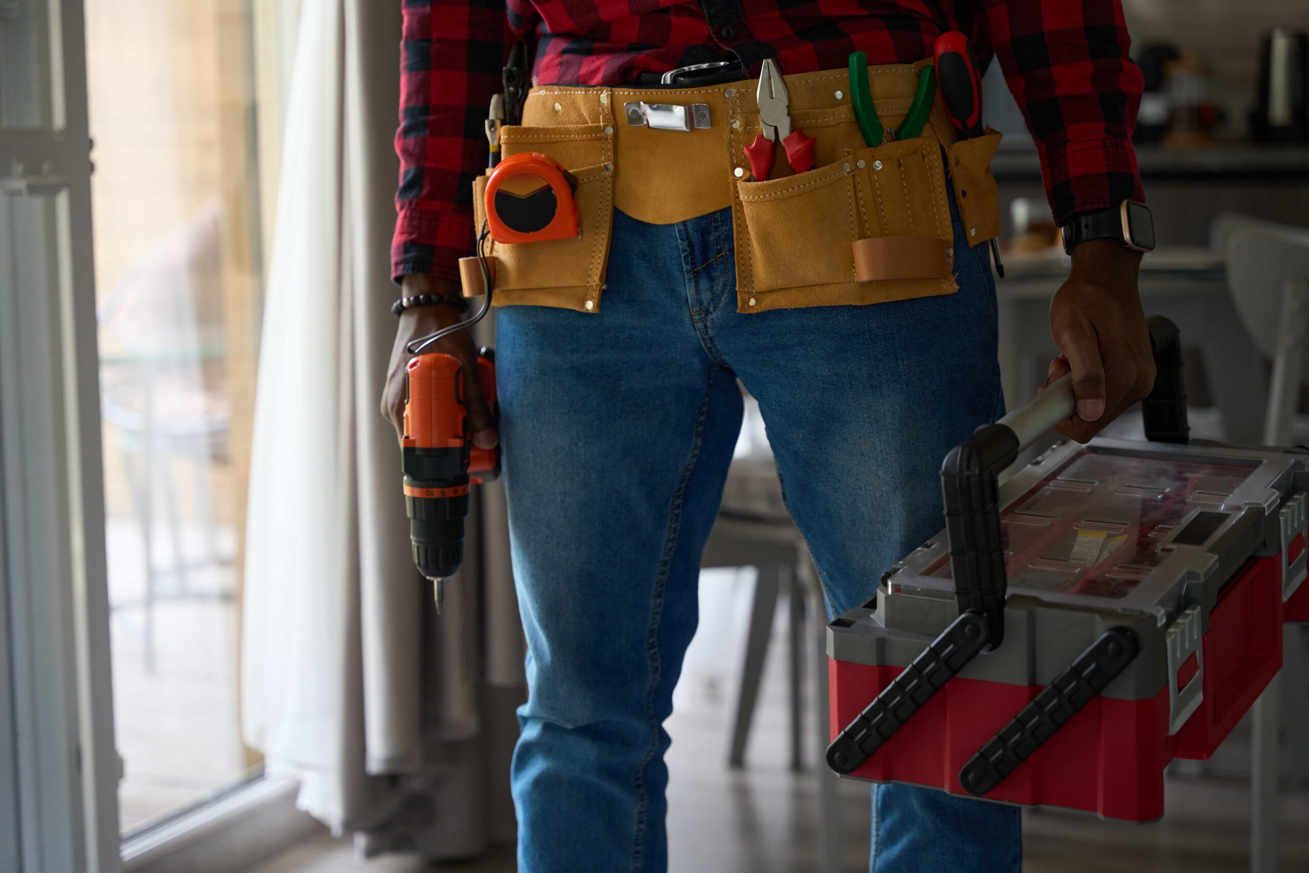 A professional handyman repair services in Atlanta, GA wearing a tool belt and holding a power drill and toolbox indoors.