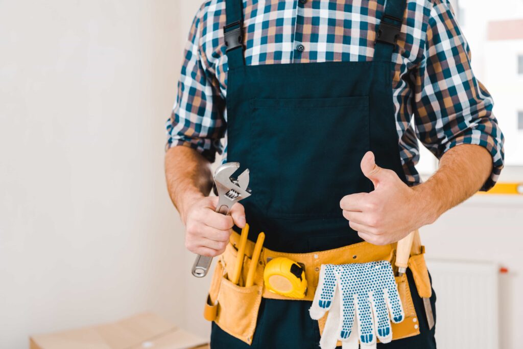 A professional handyman in Charlotte, NC wearing overalls and a tool belt, holding a wrench and giving a thumbs up.
