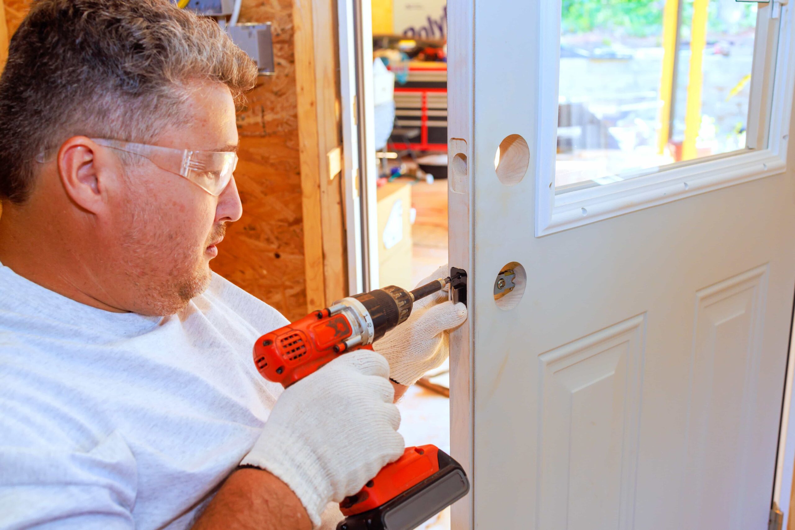 A professional locksmith wearing safety glasses and gloves uses a power drill to a Door Installation Near Me in Las Vegas, NV.