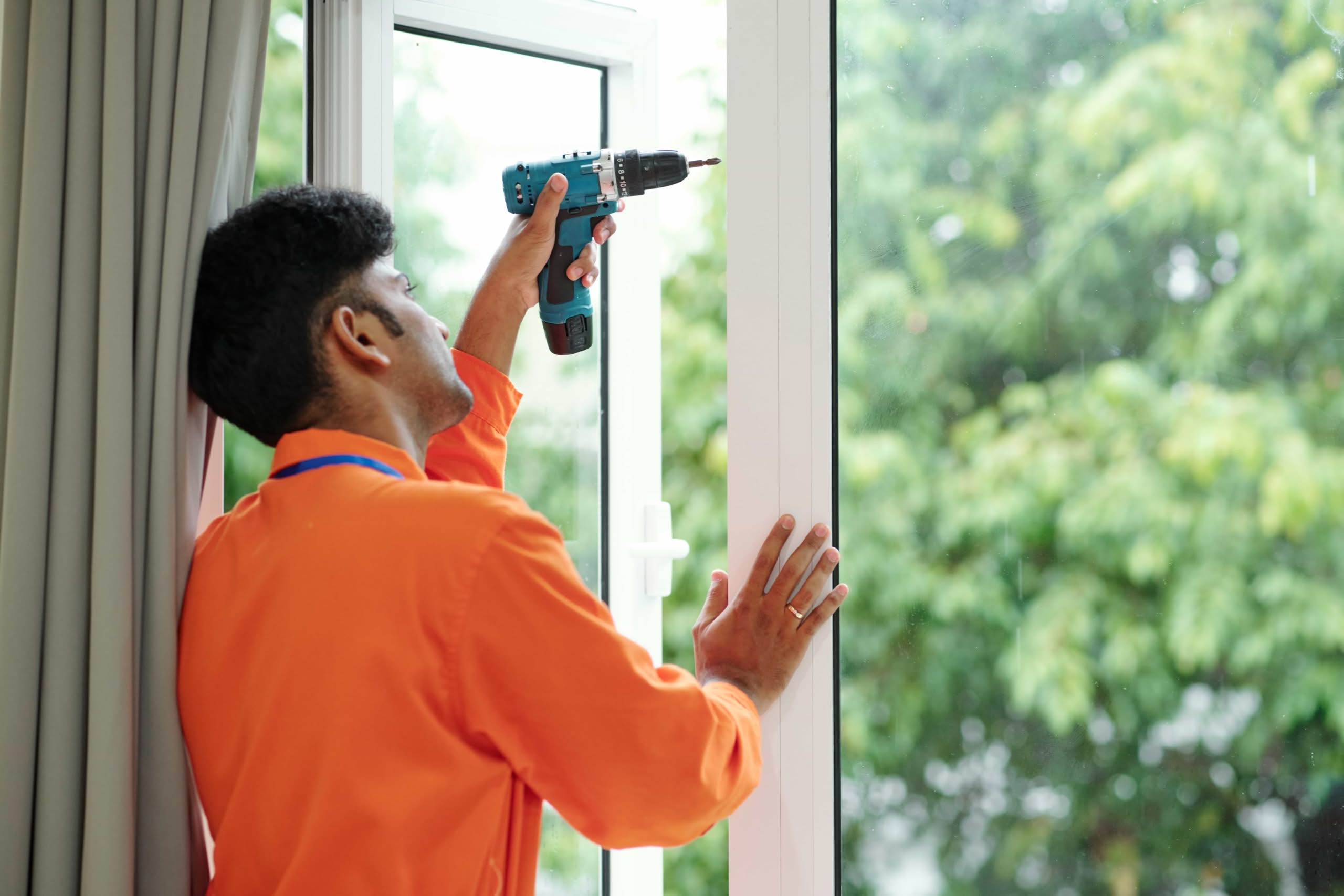 A professional technician in an orange shirt uses a power drill to secure a new impact-resistant vinyl window frame from the inside of a home to prep your windows and doors for hurricane season in Miami, Florida.